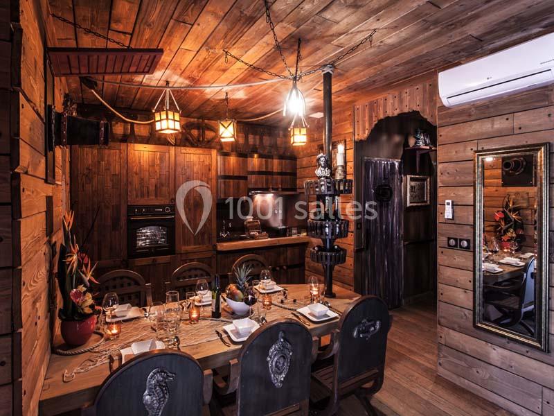 Salle à manger rustique en bois avec table dressée, cuisine ouverte et éclairage chaleureux.