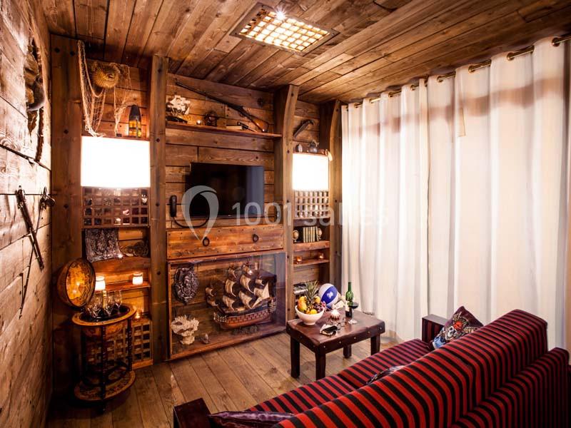 Salon chaleureux en bois avec canapé rayé, décorations maritimes, télévision murale et rideaux blancs.