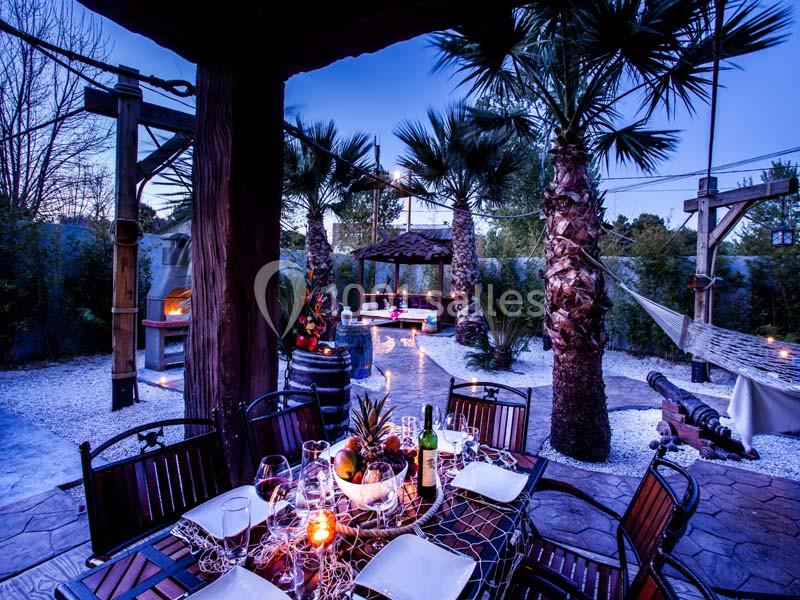 Terrasse extérieure aménagée avec table dressée, palmiers, éclairage tamisé et coin détente sous un gazebo.