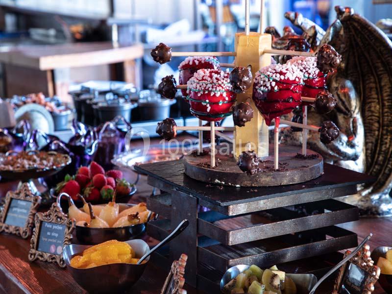 Pommes d'amour rouges décorées sur un présentoir en bois, entourées de fruits et desserts variés sur une table.