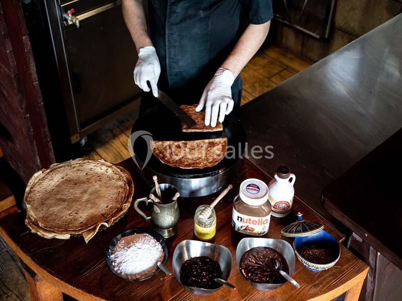 Un cuisinier prépare une crêpe sur une plaque chauffante, entouré d'ingrédients comme chocolat, miel et sucre.