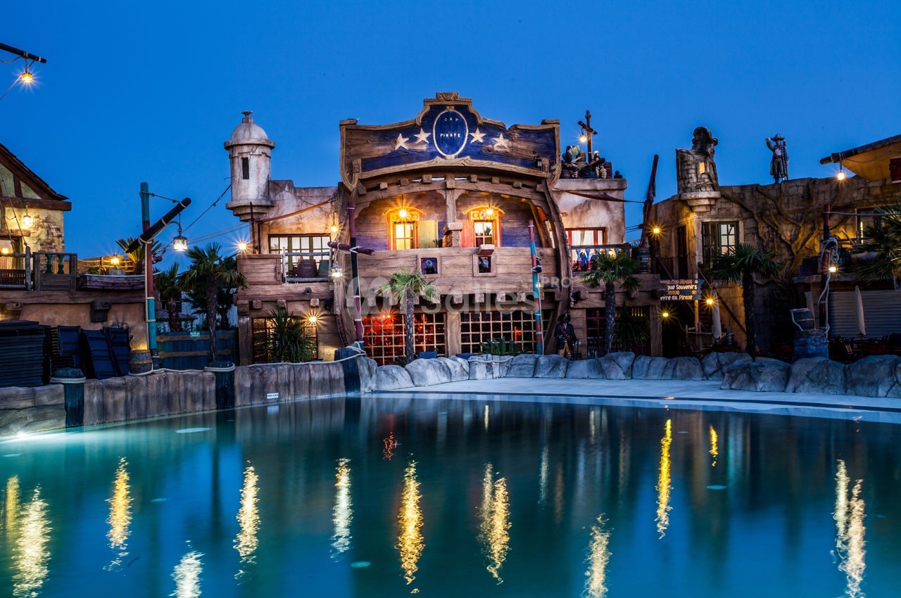 Façade d'un bâtiment à thème pirate éclairé la nuit, avec une piscine calme au premier plan.