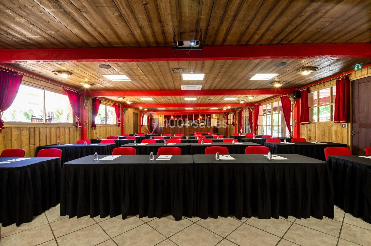 Salle de conférence avec des tables disposées en rangées, nappées de noir, et des chaises rouges, éclairée par la lumière…