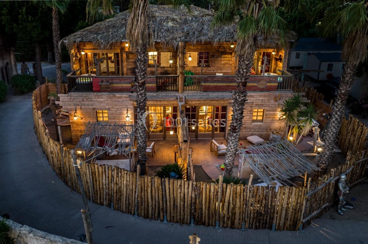 Maison en bois à deux étages avec toit en chaume, entourée de palmiers et d'une clôture en bois, éclairée en soirée.