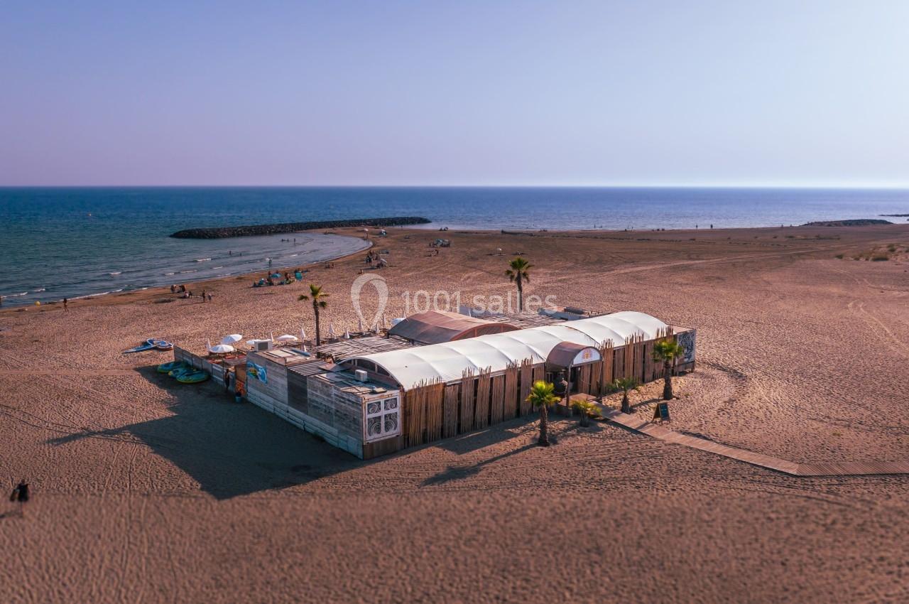 Vue aérienne d'une plage avec un bâtiment en bois et toile, entouré de sable, près de la mer calme.