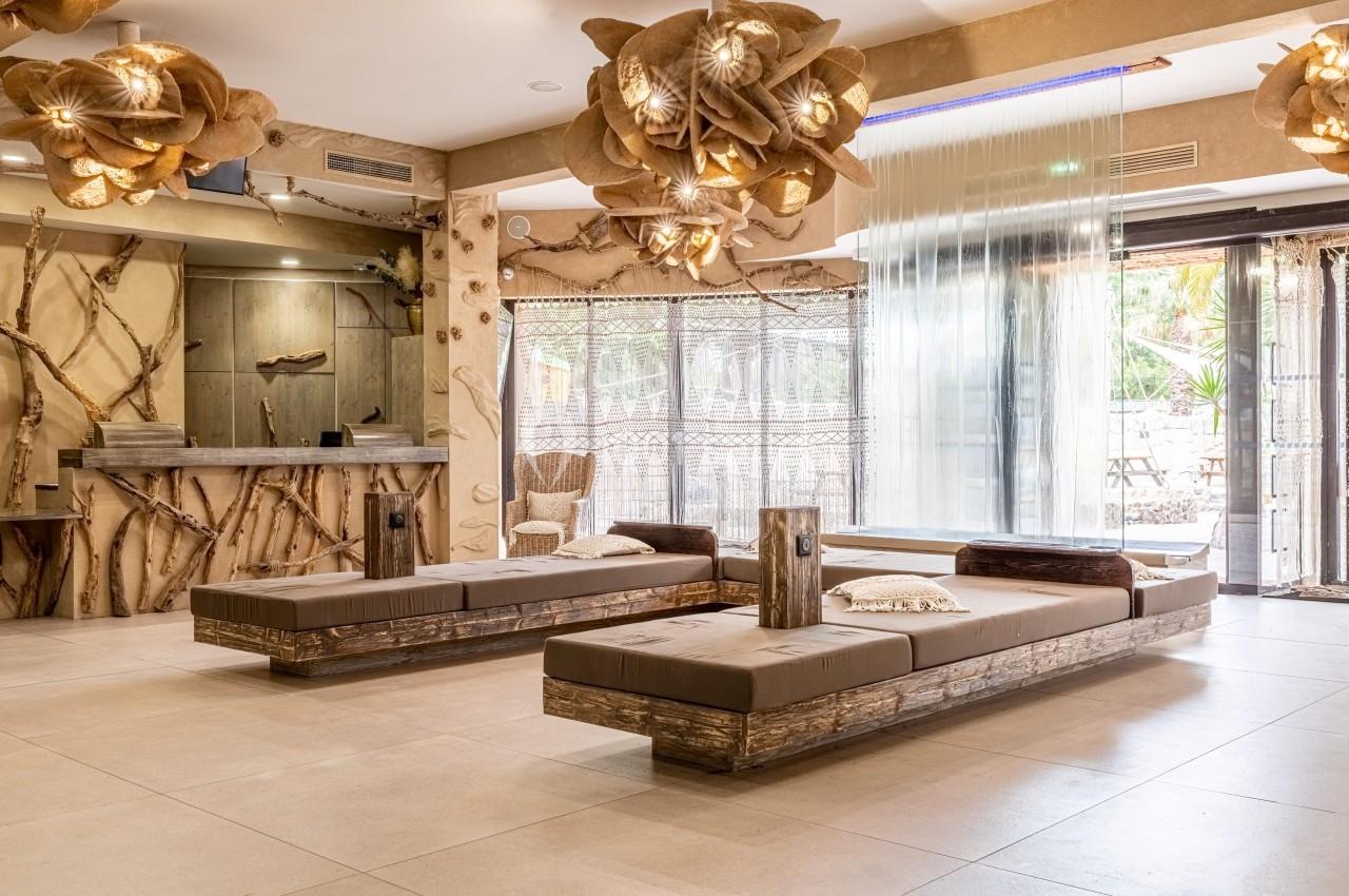 Salon moderne avec banquettes en bois, décor naturel, luminaires suspendus et grande baie vitrée donnant sur l'extérieur.