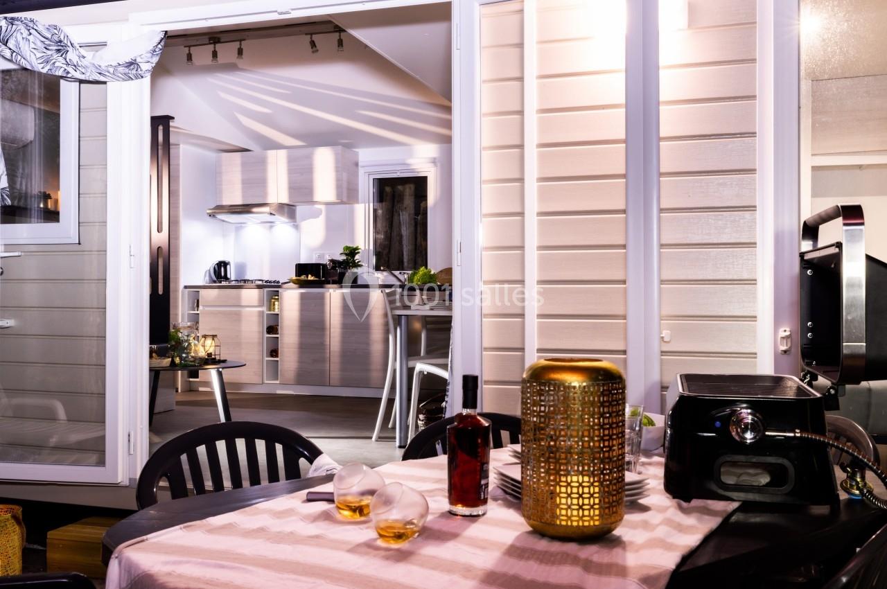 Vue d'une terrasse aménagée avec table dressée, donnant sur une cuisine moderne éclairée à l'intérieur.