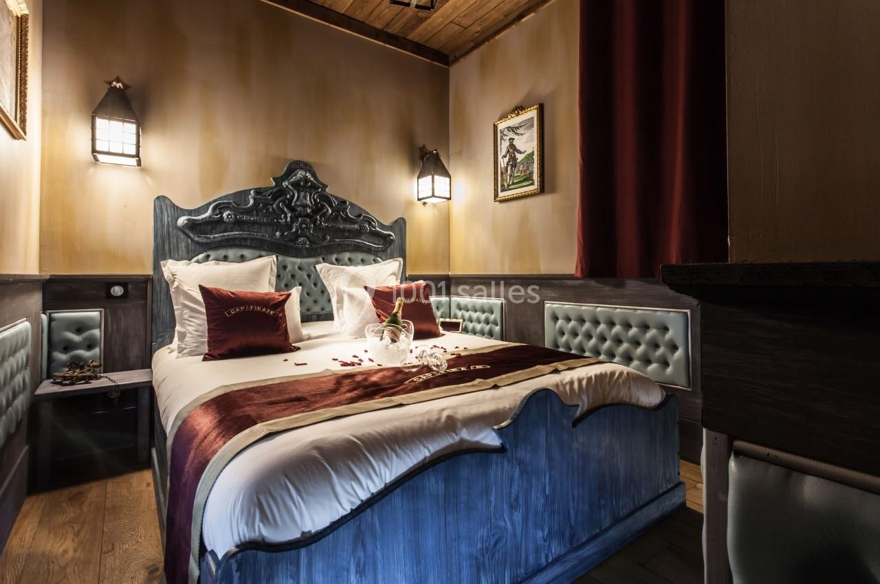 Chambre décorée avec un lit double, draps blancs, coussins rouges, éclairage tamisé et détails en bois et cuir capitonné.