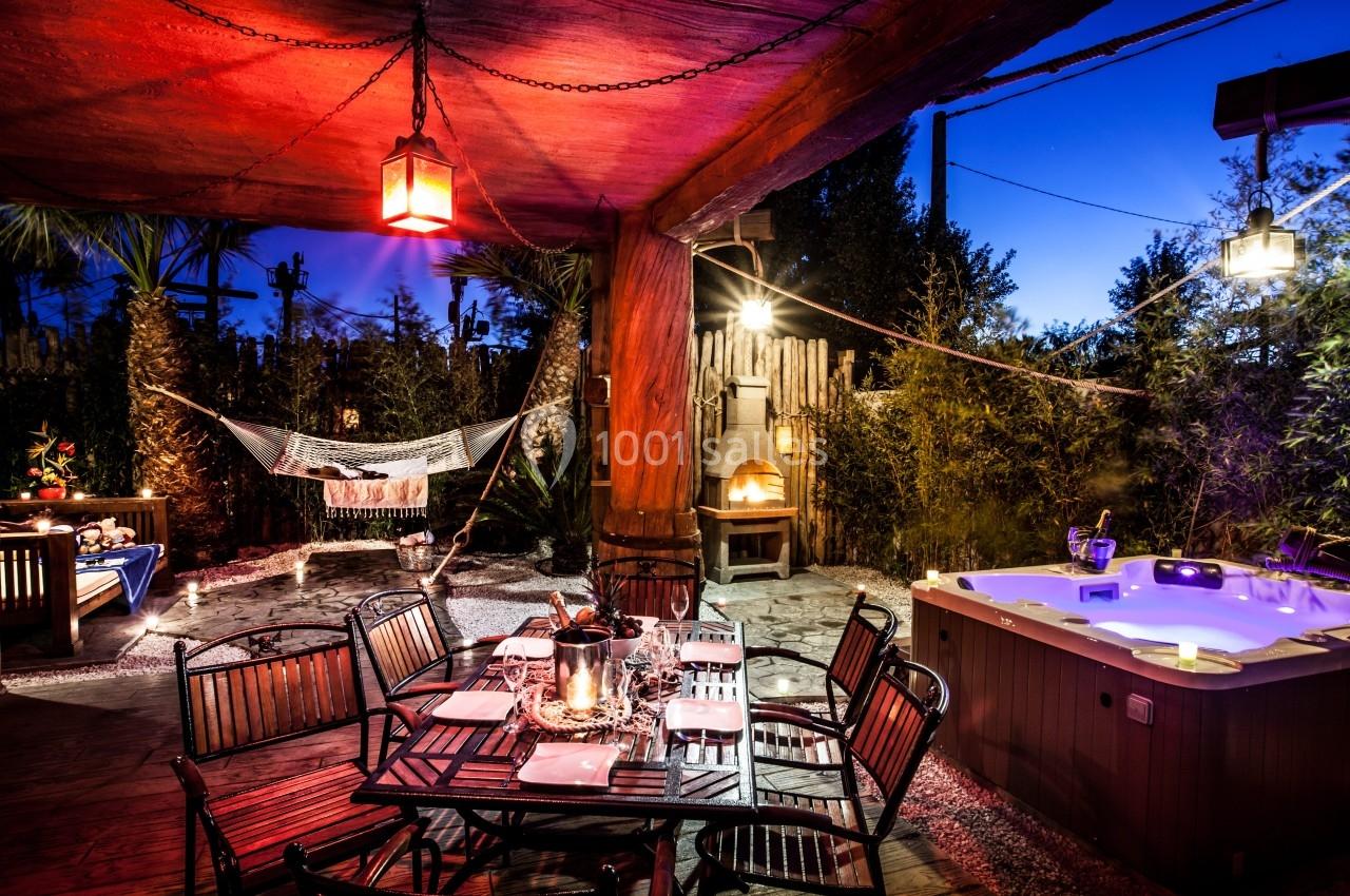 Terrasse éclairée de nuit avec table dressée, jacuzzi allumé, hamac suspendu et four extérieur en arrière-plan.