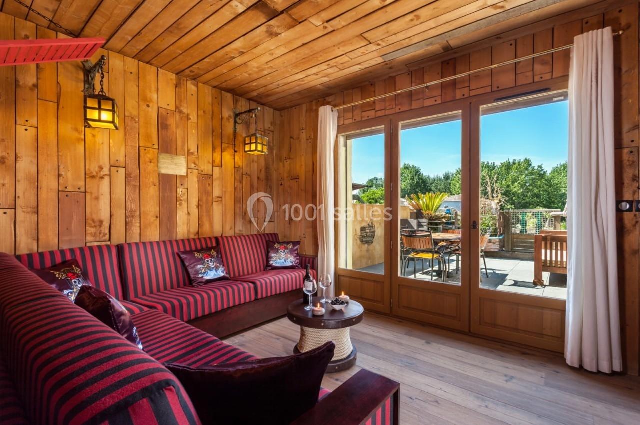Salon avec murs en bois, canapé rayé rouge et noir, table basse, et grande baie vitrée donnant sur une terrasse ensoleillée.