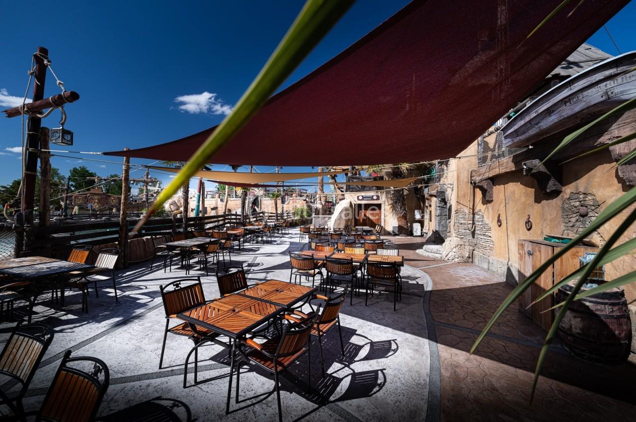 Terrasse extérieure avec tables et chaises en bois sous des voiles d'ombrage, entourée d'une décoration thématique.