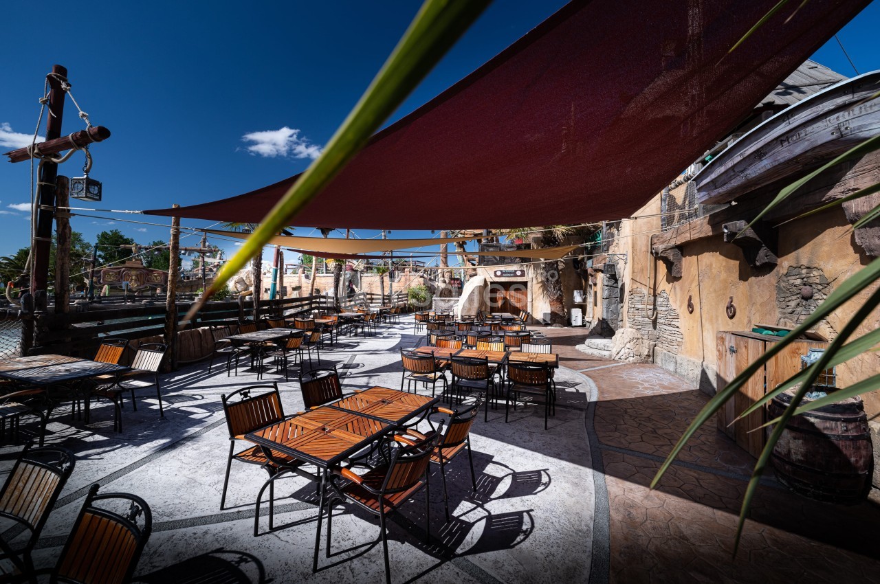 Terrasse extérieure avec tables et chaises en bois sous des voiles d'ombrage, entourée d'une décoration thématique.