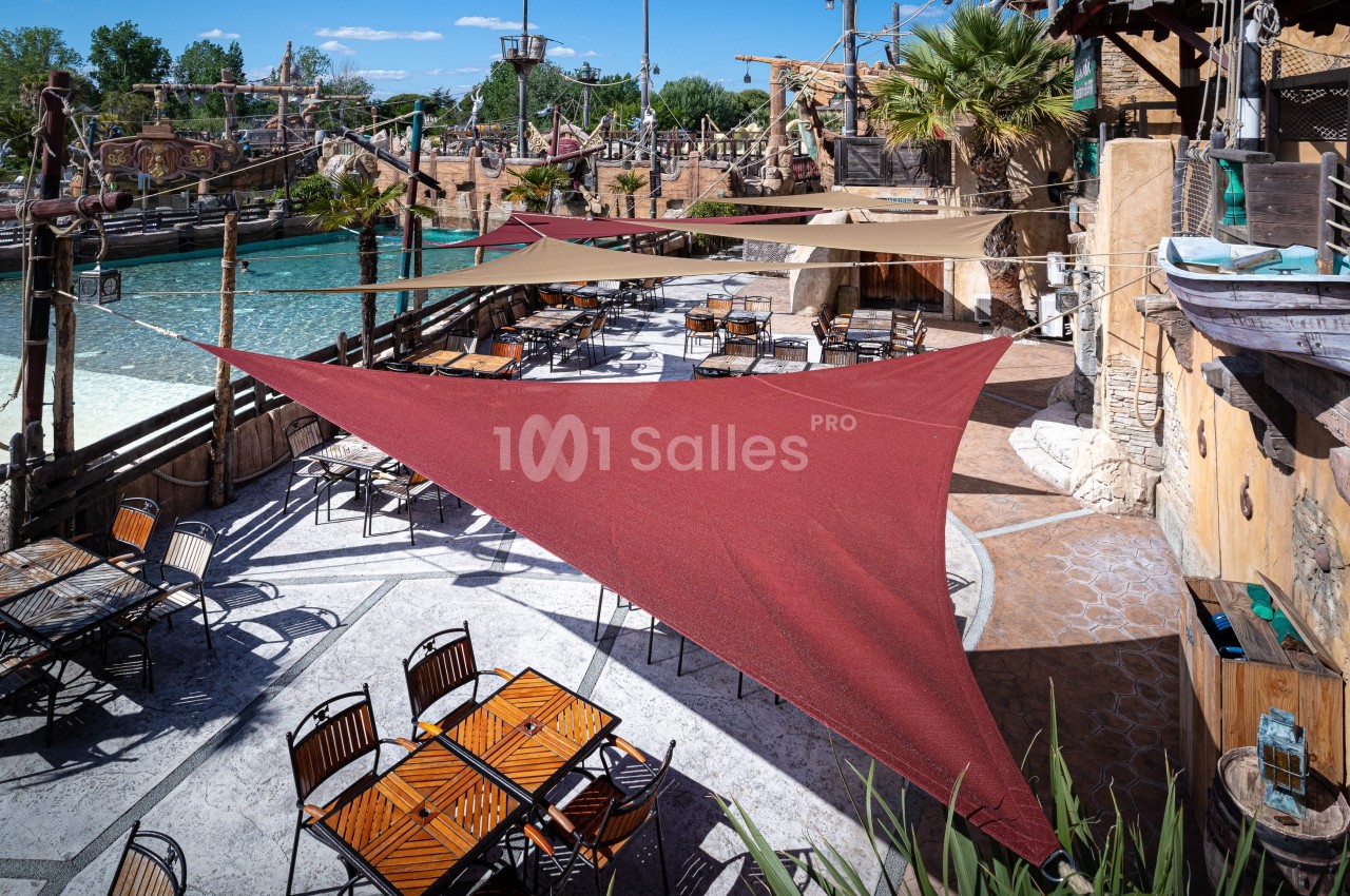 Terrasse extérieure avec tables et chaises sous des voiles d'ombrage rouges, près d'un décor nautique et d'une étendue d'eau.