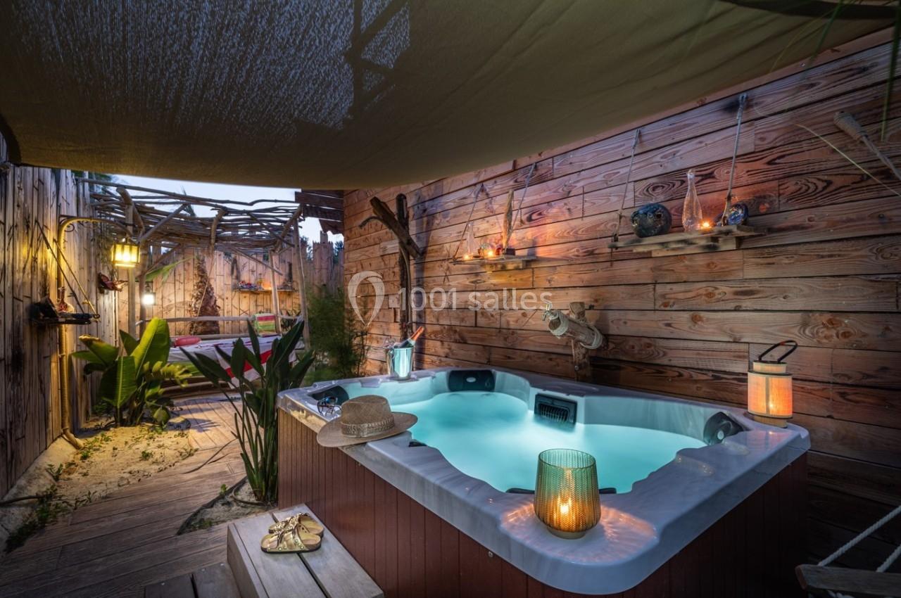 Jacuzzi extérieur éclairé le soir, entouré de bois et de plantes, avec des lanternes et une ambiance chaleureuse.
