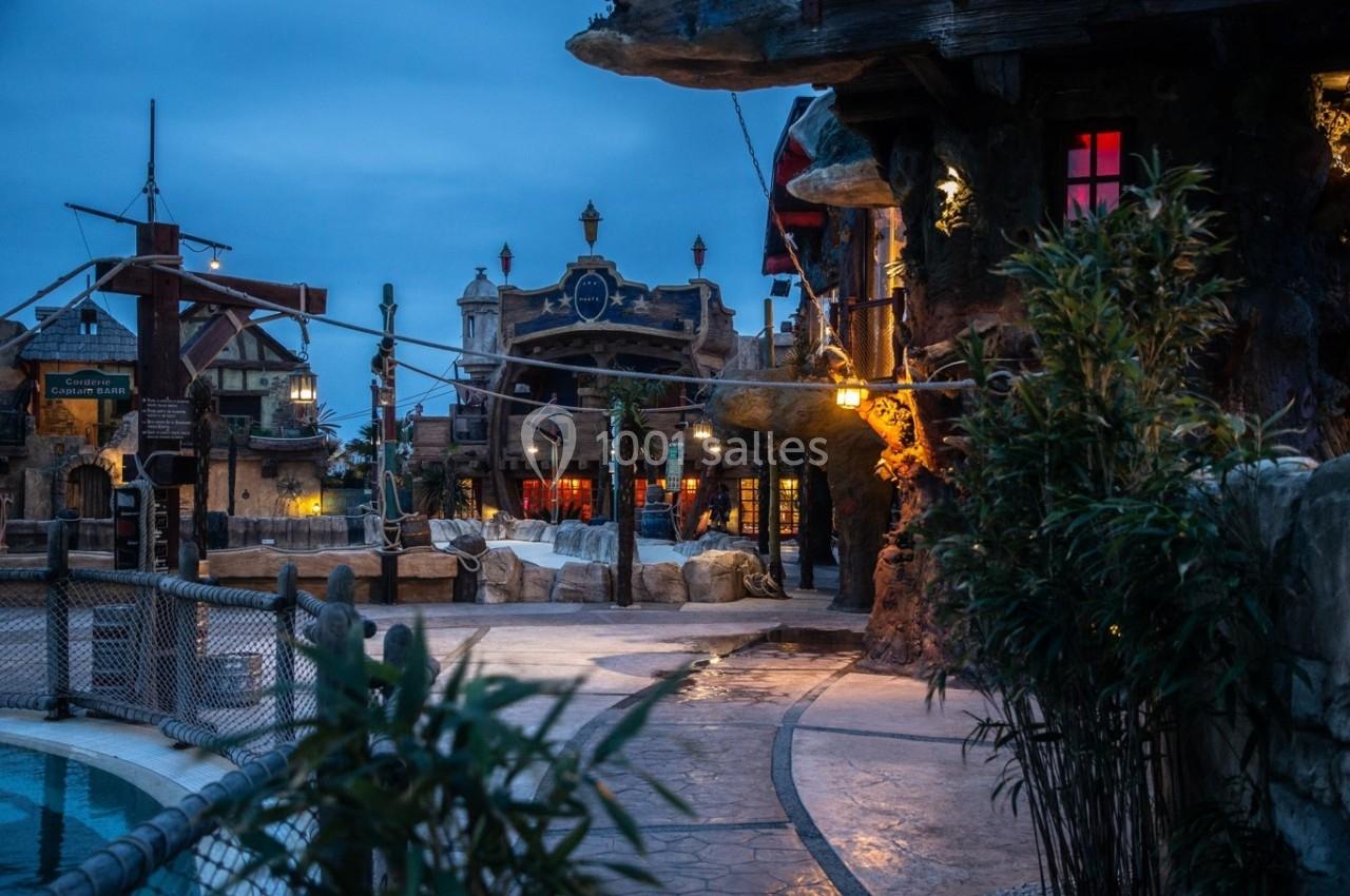 Village thématique au crépuscule avec bâtiments éclairés, décorations maritimes et végétation en premier plan.