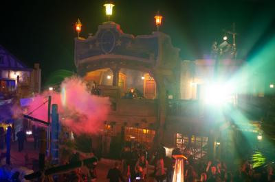 Vue nocturne d'un complexe de loisirs avec piscine illuminée, bâtiments thématiques et paysage urbain en arrière-plan.