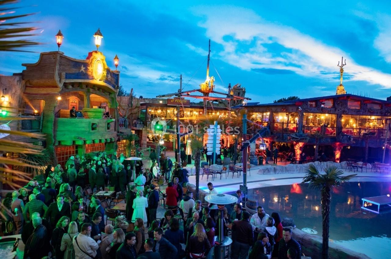 Foule rassemblée dans un lieu extérieur animé, décoré sur le thème des pirates, avec éclairages colorés et piscine.