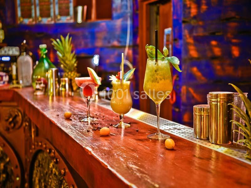Trois cocktails colorés servis sur un comptoir en bois dans un bar à l'ambiance chaleureuse et exotique.