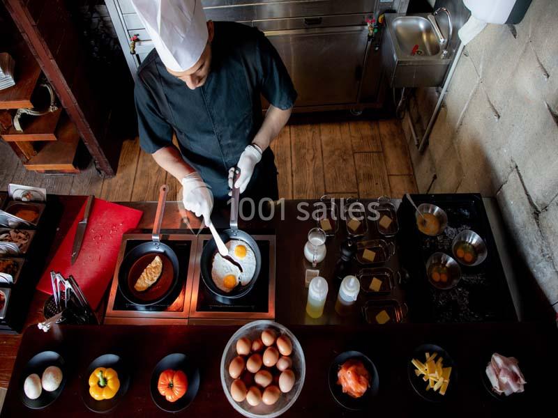 Un chef en tenue noire prépare des œufs au plat dans une cuisine professionnelle avec divers ingrédients disposés.