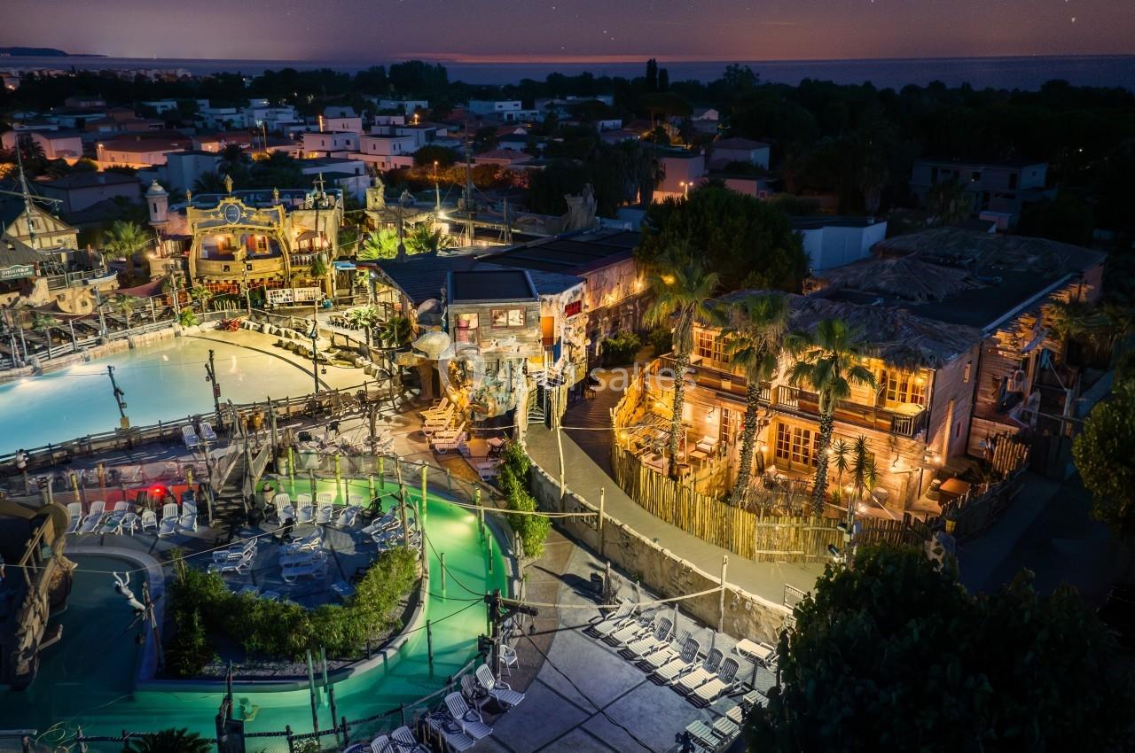 Vue nocturne d'un complexe de loisirs avec piscine éclairée, bâtiments thématiques et habitations en arrière-plan.