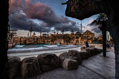 Vue nocturne d'un complexe de loisirs avec piscine illuminée, bâtiments thématiques et paysage urbain en arrière-plan.