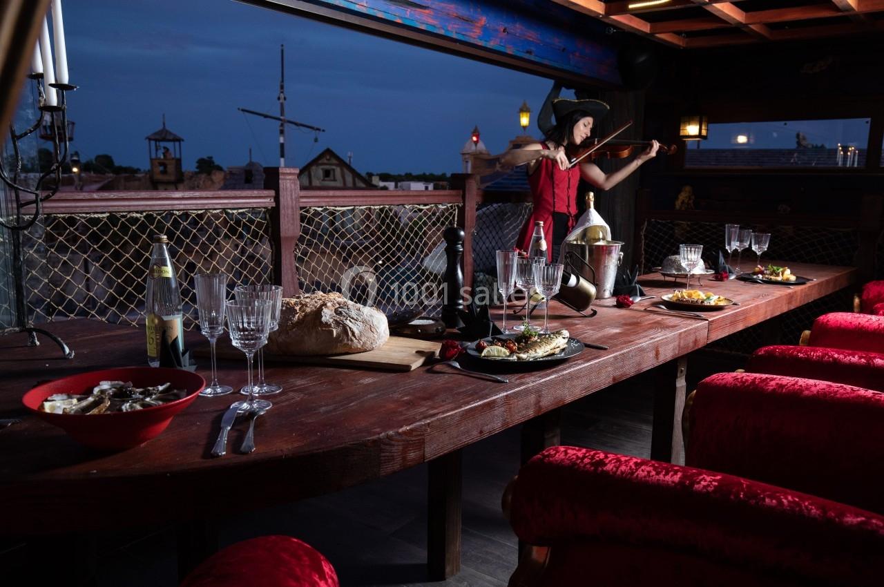 Table en bois dressée avec des plats et boissons, vue nocturne sur un décor extérieur, femme jouant du violon en arrière…