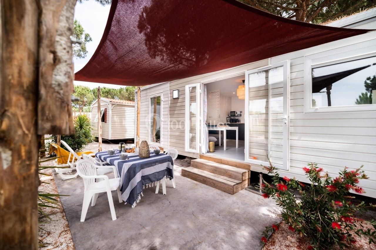 Terrasse d'un mobil-home avec table dressée, chaises blanches, voile d'ombrage rouge et végétation environnante.