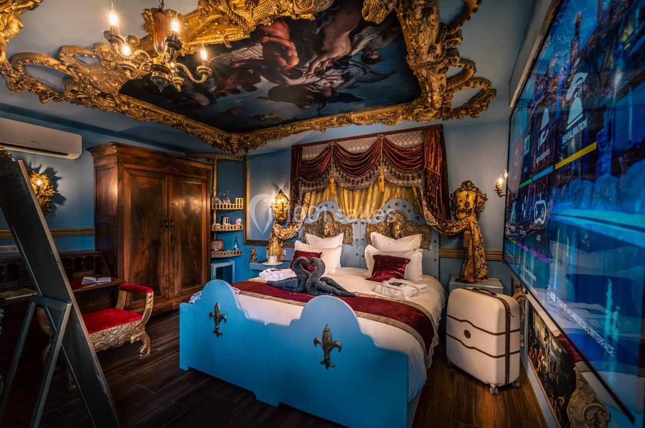 Chambre décorée de manière baroque avec lit bleu, fresque au plafond, meubles anciens et éclairage chaleureux.
