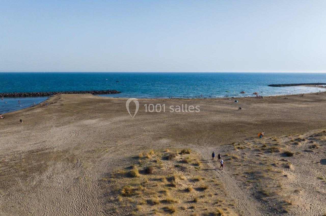 Vue aérienne d'une plage de sable avec quelques promeneurs, des dunes au premier plan et la mer en arrière-plan.