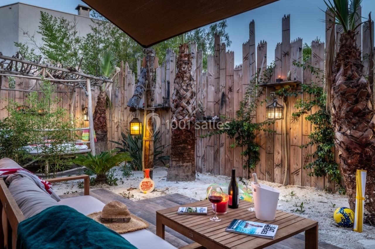 Terrasse aménagée avec canapé, table basse, palmiers, éclairage chaleureux et décor en bois dans un jardin extérieur.