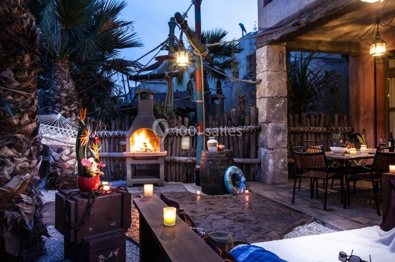 Terrasse extérieure avec cheminée allumée, table dressée, plantes tropicales et éclairage tamisé en soirée.
