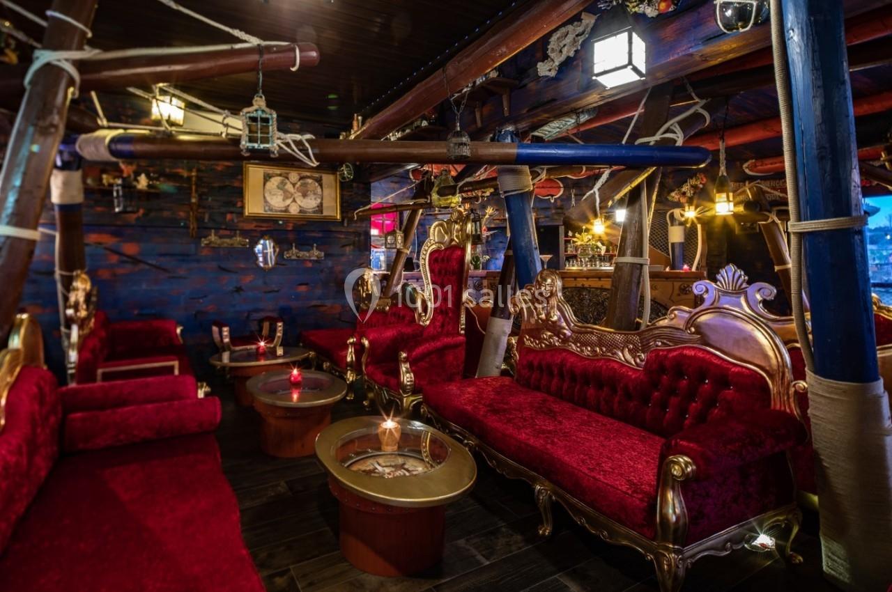 Salon intérieur avec canapés rouges ornés, tables basses en bois et décorations maritimes dans une ambiance tamisée.