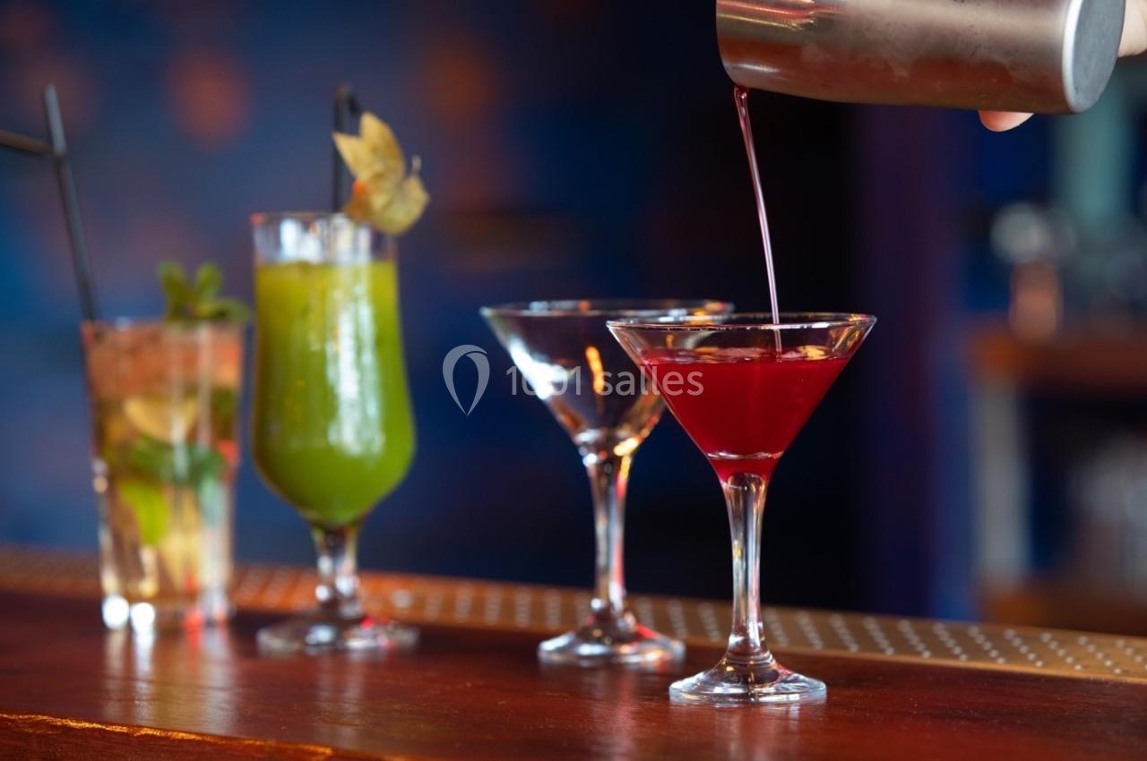 Quatre cocktails colorés alignés sur un bar, dont un rouge servi depuis un shaker en métal.