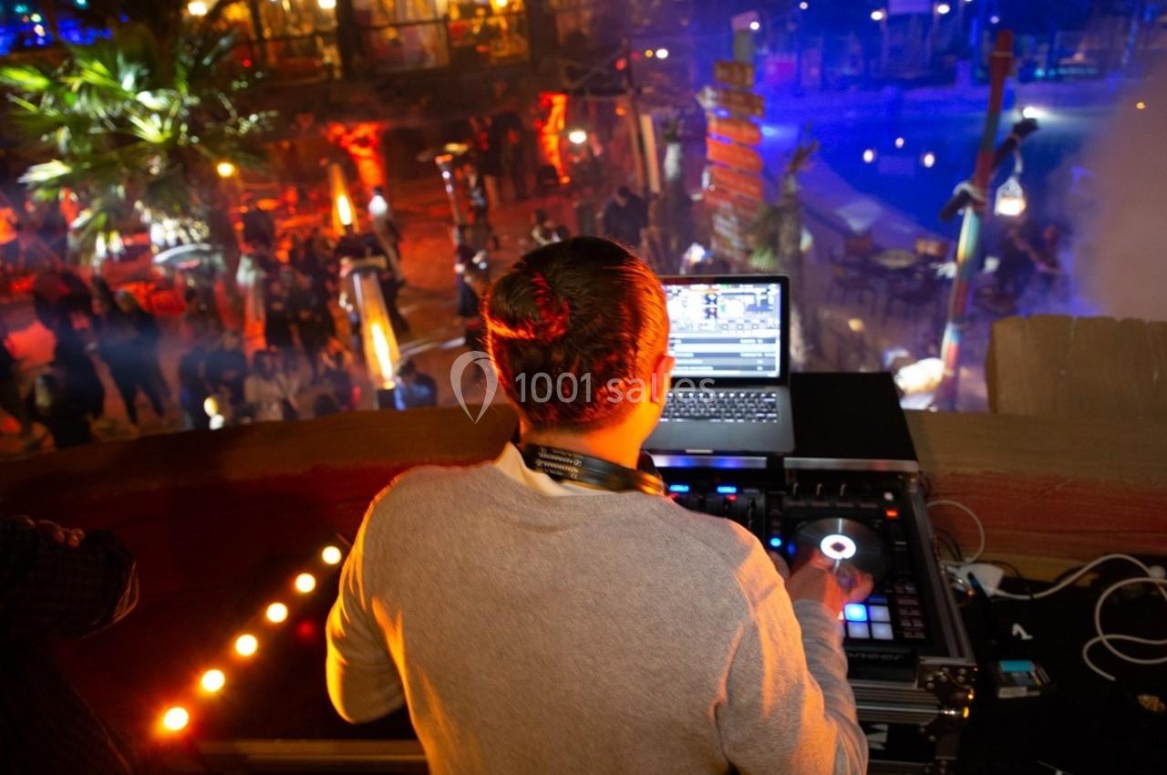 Un DJ mixe de la musique sur une console, face à une foule dans un lieu animé et éclairé par des lumières colorées.