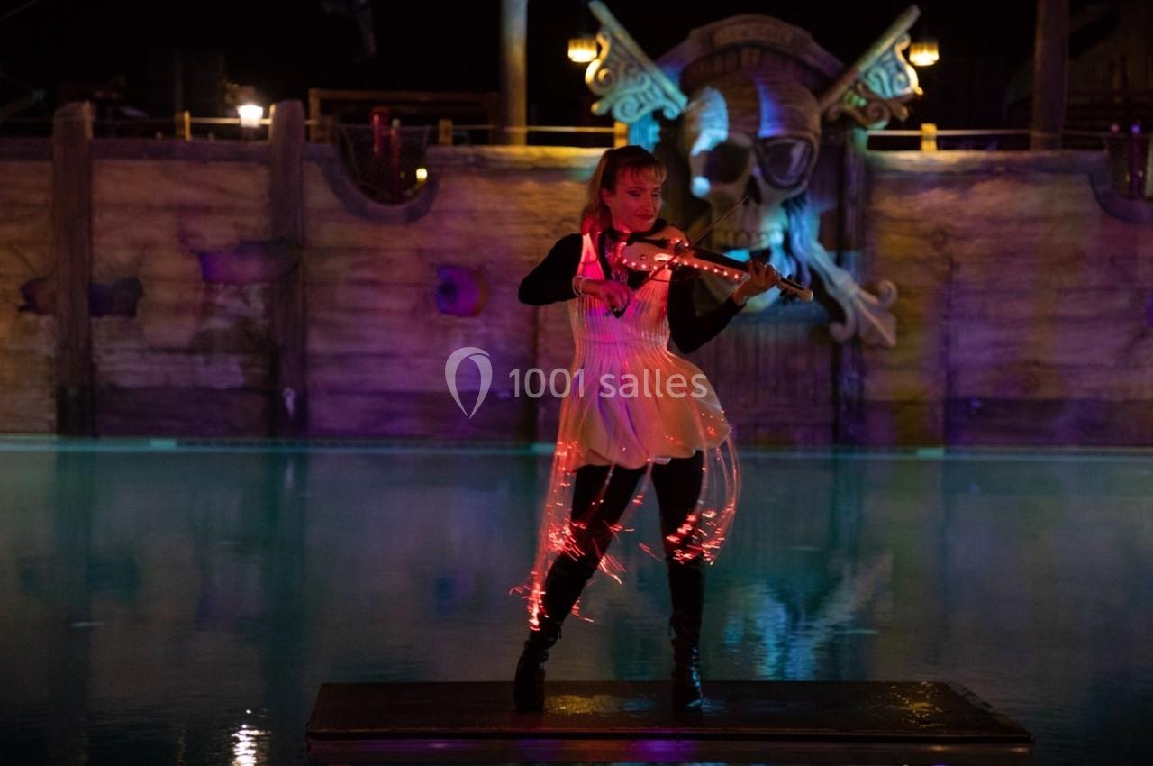 Une violoniste joue sur une scène flottante éclairée, devant un décor de bateau pirate illuminé.