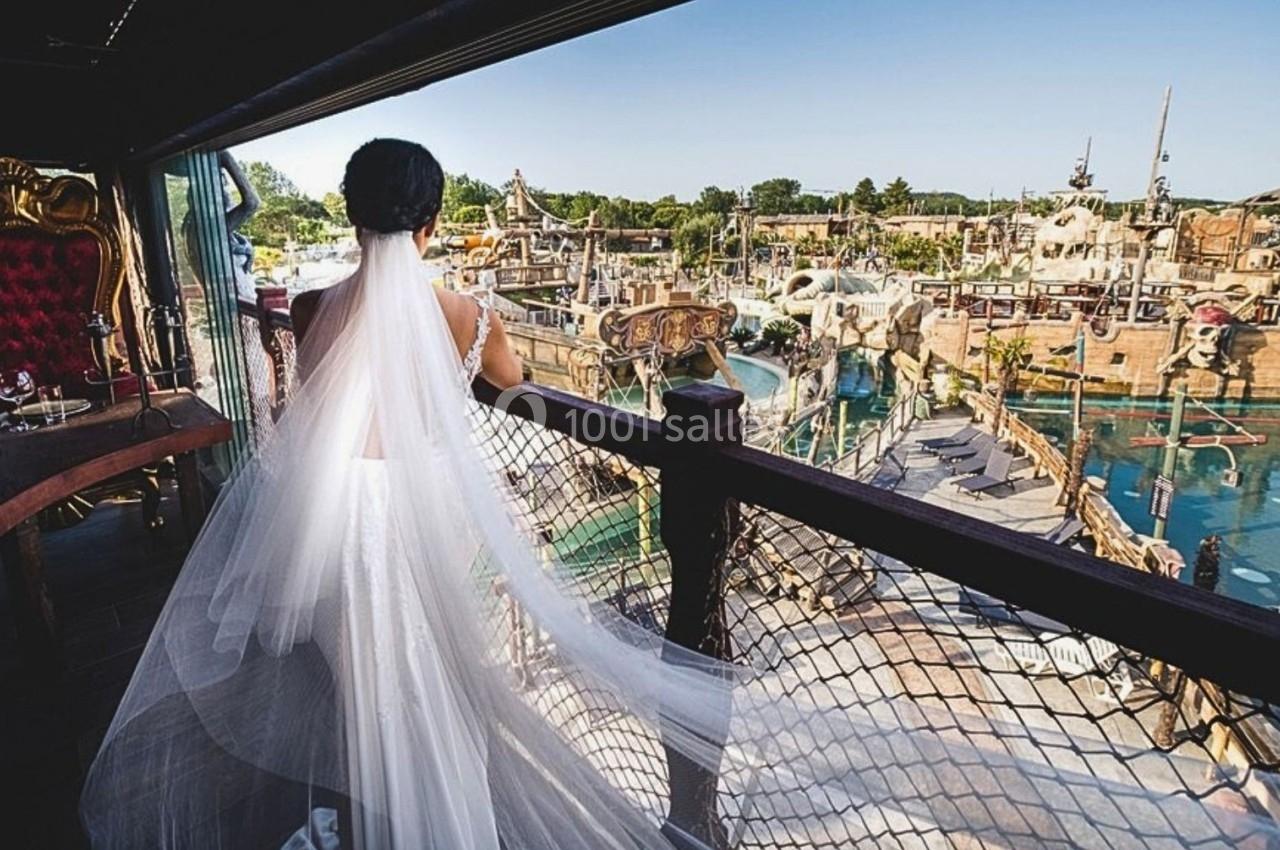 Une mariée de dos, en robe blanche avec un long voile, observe un parc aquatique depuis une terrasse en bois.