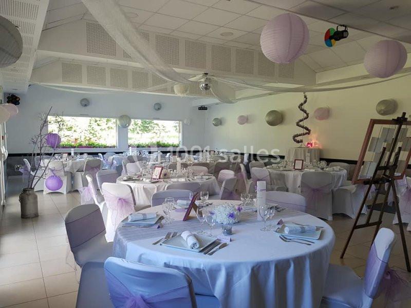 Salle de réception décorée avec des tables rondes, nappes blanches, chaises ornées de rubans mauves et lanternes suspendues.