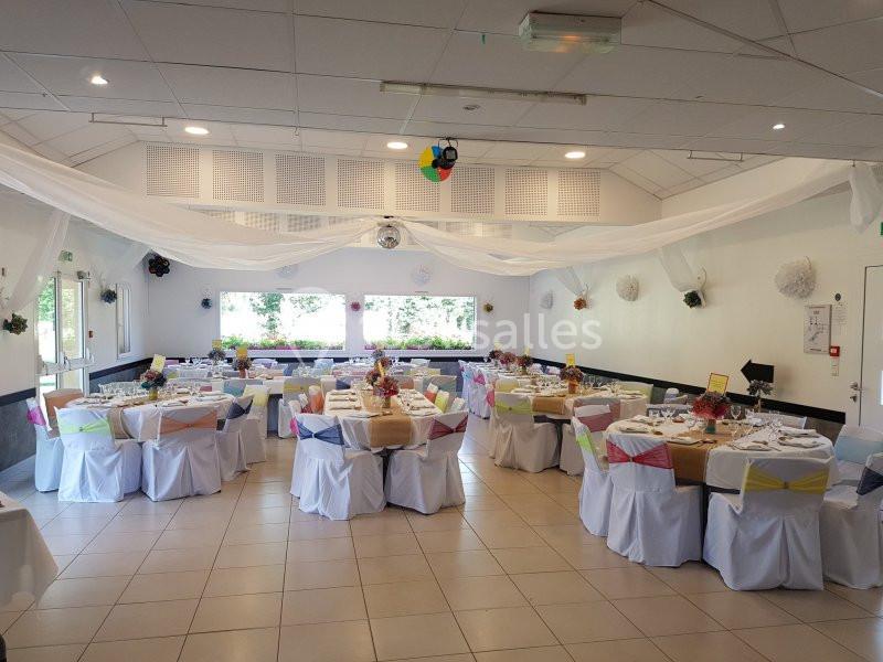 Salle de réception décorée avec des tables dressées, nappes blanches et chaises ornées de rubans colorés.