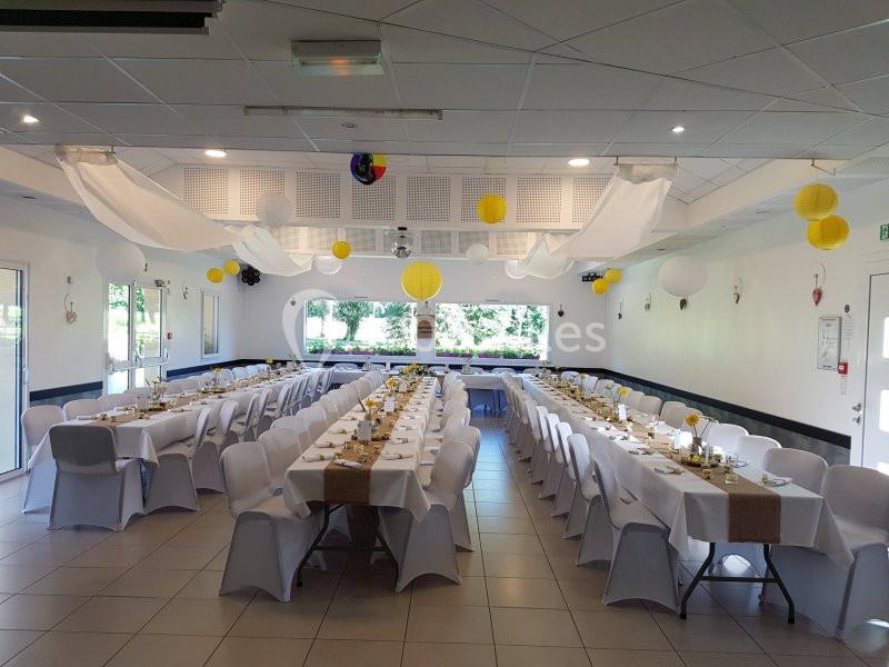 Salle décorée pour un événement, avec longues tables dressées, nappes blanches et lanternes jaunes suspendues.