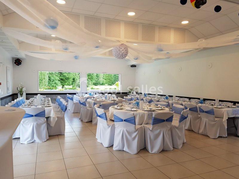 Salle de réception décorée avec des tables dressées, nappes blanches, rubans bleus et vue sur un jardin verdoyant.