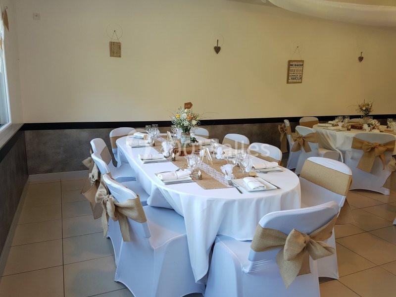 Salle décorée pour un événement, avec des tables rondes dressées, nappes blanches et chaises ornées de nœuds en toile de…