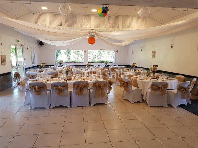 Salle de réception décorée avec des tables rondes dressées, chaises ornées de nœuds beiges et éclairage naturel.