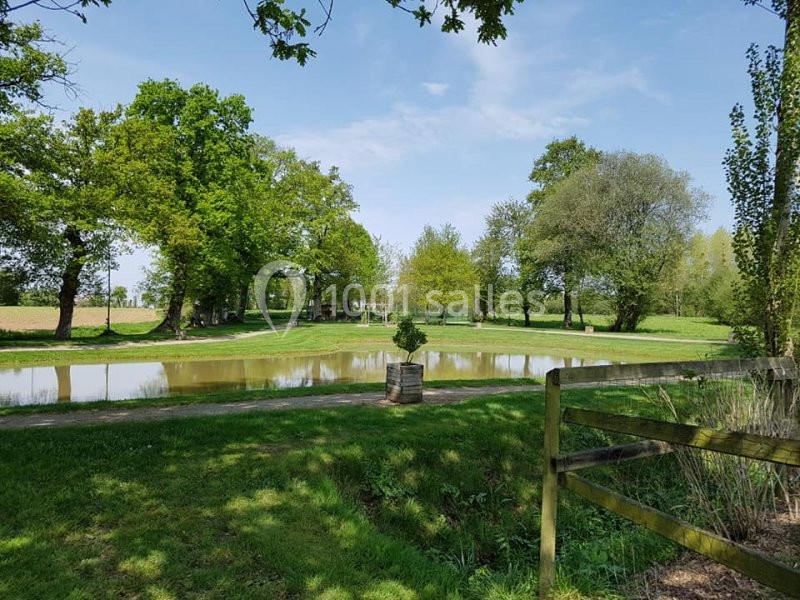 Paysage champêtre avec un étang entouré d'arbres, une barrière en bois au premier plan et un ciel dégagé.