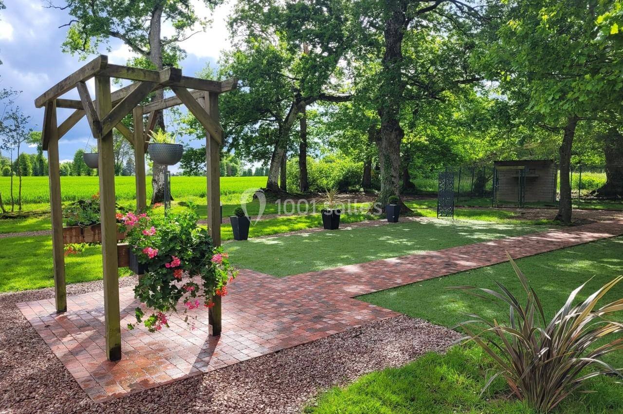 Jardin aménagé avec pergola en bois, allée pavée, pelouse artificielle et pots de fleurs, entouré d'arbres.