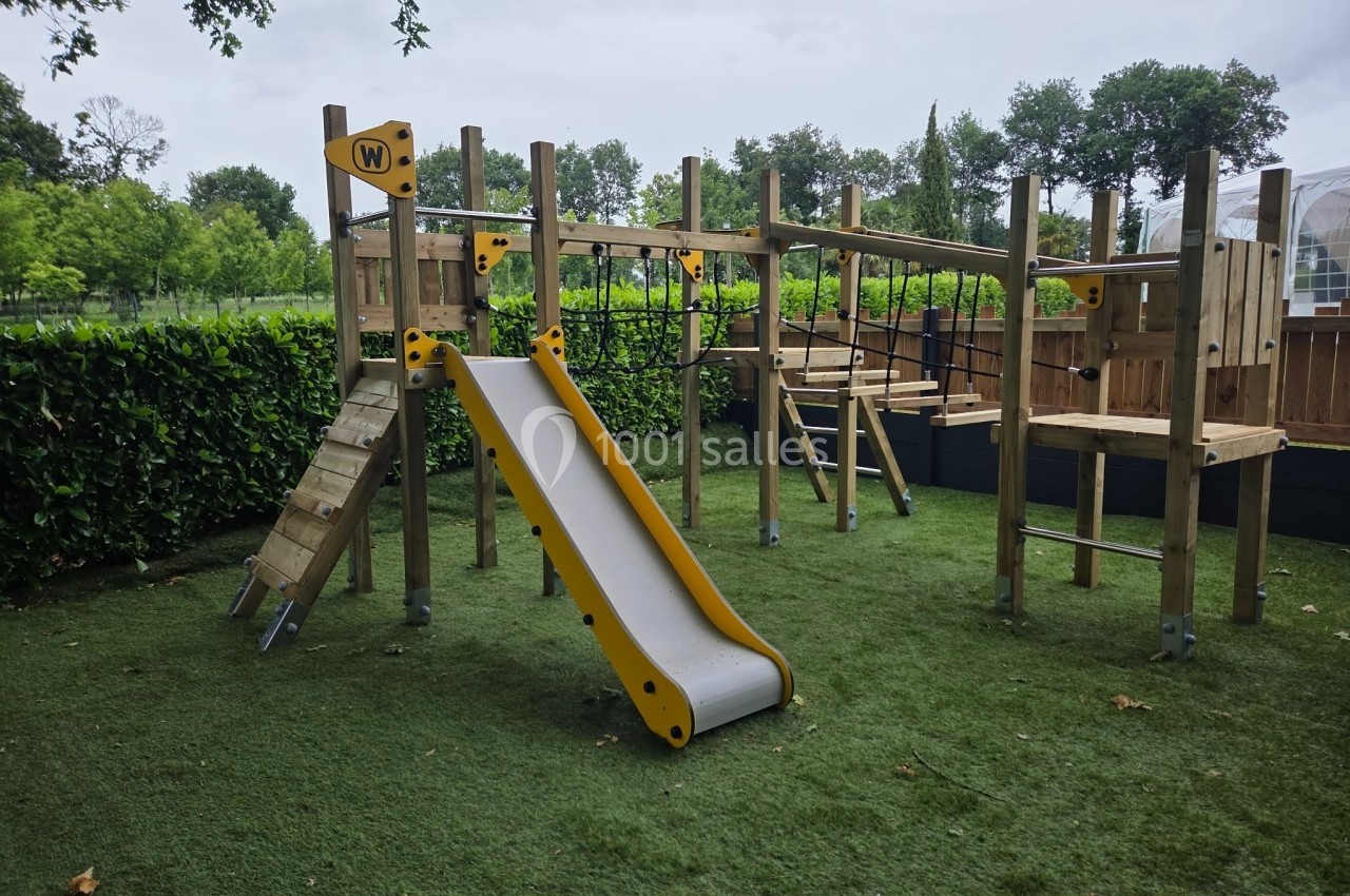Structure de jeu en bois avec toboggan, pont suspendu et filets, installée sur une pelouse artificielle entourée de haies.