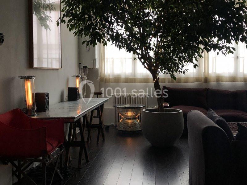 Espace intérieur lumineux avec un grand arbre en pot, un bureau en marbre, des fauteuils et un canapé sombre.