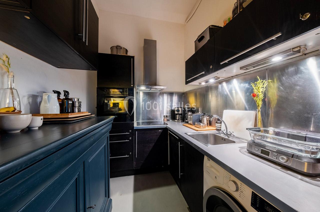 Cuisine moderne avec meubles noirs, plan de travail en bois, électroménagers et crédence métallique réfléchissante.