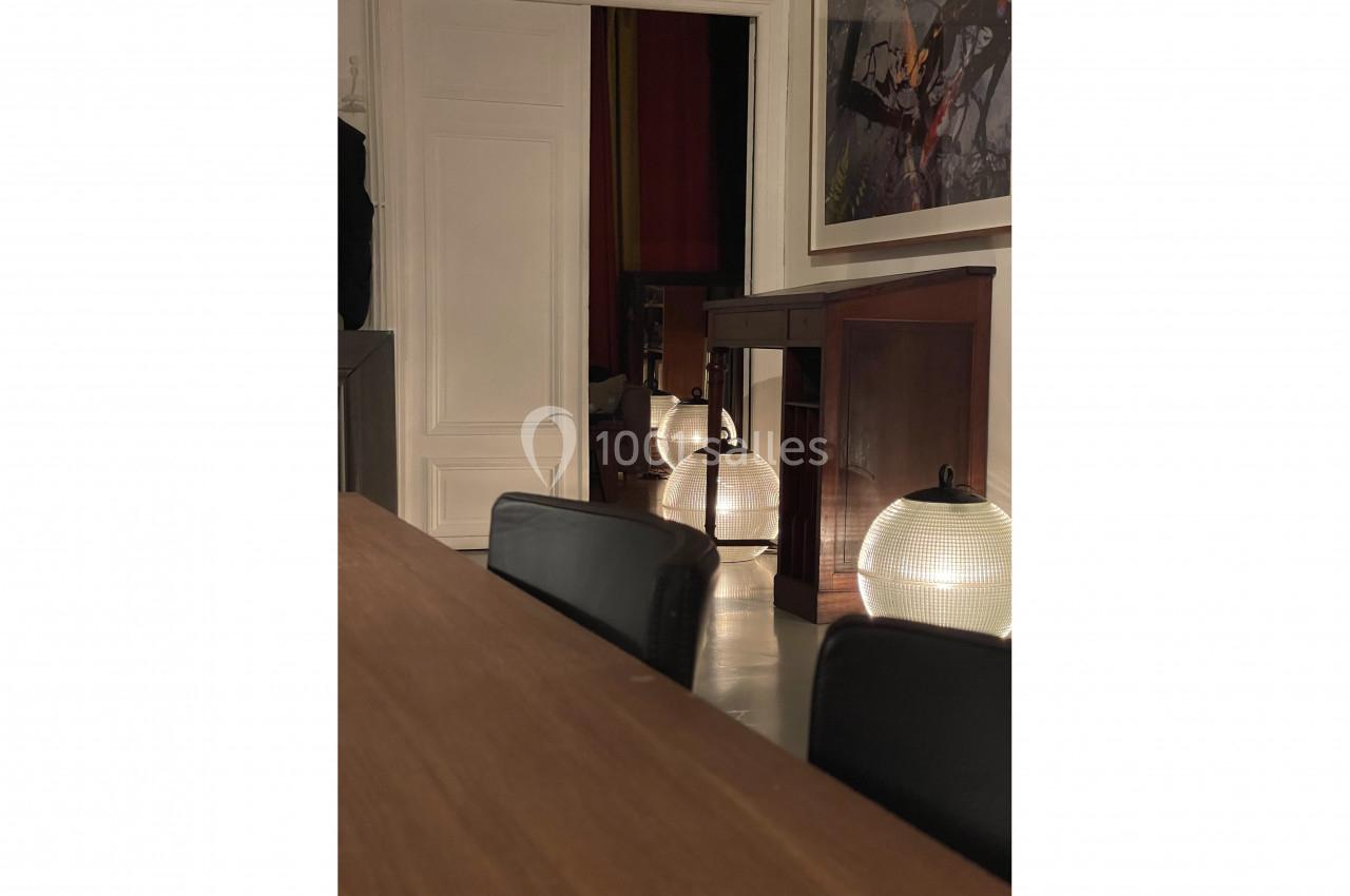 Vue intérieure d'une pièce avec table en bois, chaises noires et lampes rondes éclairant un couloir en arrière-plan.