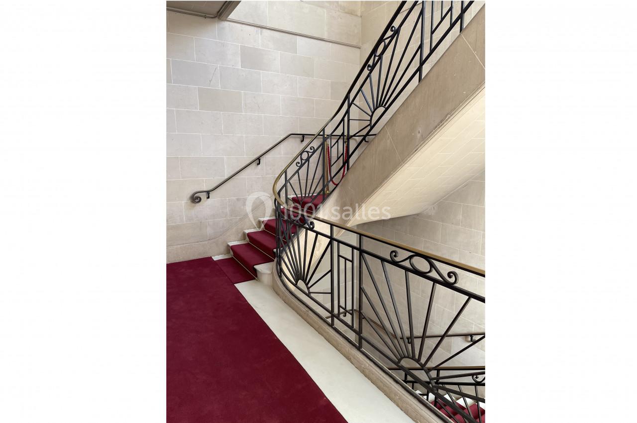 Escalier en pierre avec tapis rouge et rampe en fer forgé, dans un intérieur lumineux aux murs clairs.