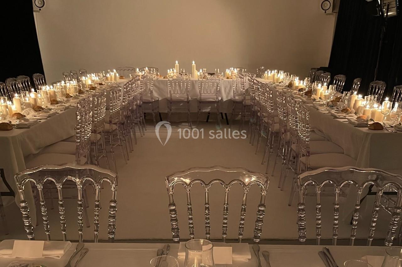 Salle de réception avec tables en U décorées de bougies, chaises transparentes et vaisselle élégante.